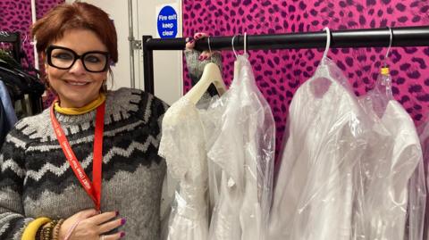 Vivienne Wood has short red hair, is wearing large thick-rimmed black glasses and a grey and black sweater. She has a red lanyard draped over her neck. She is standing beside a rail of four white First Communion dresses. The room she is standing in has pink walls and is covered in black spots 