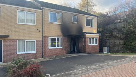 The fire-stained front of an end-of-terrace house. Black fire damage is above the front door.
