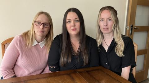 Three women sat at a kitchen table in a row. They are looking at the camera and have serious looks on their faces.
