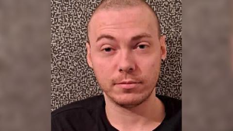 Grant McKinnon is looking straight at the camera. He has a shaved head and is wearing a black top