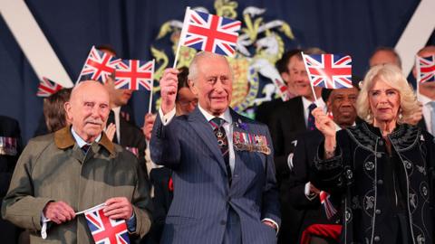 King Charles and Queen Camilla waving flags next to 107-year-old World War Two bomber pilot Harry Richardson.
