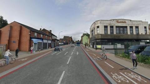 Artist impression of the main route through Attercliffe complete with red route lines, separate red cycle lanes on both sides of the carriageway, and a main road mainly free of traffic, except for two cars and a blue bus in the distance. The illustration features three cyclists, shop fronts (many of which are closed) and the former Burton building. 