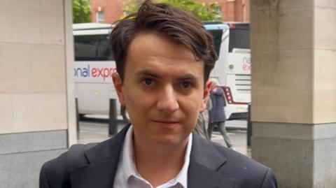 Gabriel Leroy pictured staring straight into the camera, wearing an open neck shirt and a grey/blue jacket
