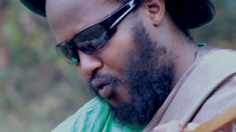 Close up of Karasira's face. He has a beard, is wearing sunglasses and is holding a musical instrument, just slightly visible in the bottom left corner. the rim of a hat and also be seen on either side of his head. His lips are pursed as he is singing