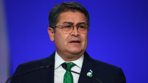 Honduras President Juan Orlando Hernández speaks in front of a blue backdrop. He has short hair and wears square-framed eye glasses. 