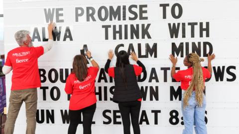 Four people, placing large black letters on a white board with their back to the camera