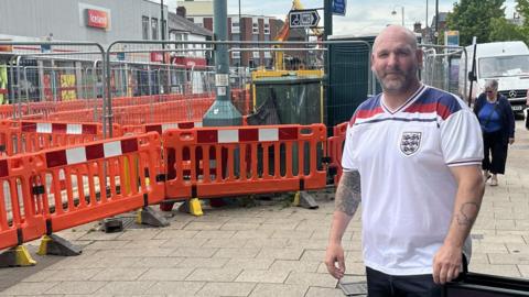 Roy Barrett a bald man with a white blue and red England shirt on and tattoos on his arms. He is stood outside his pub with the roadworks visible just behind him.