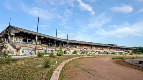 A derelict and unused speedway course, with grass and moss on the tracks. The stands have graffiti on them.