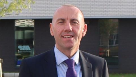 A head and shoulders shot of a man with a shaved head standing outdoors, smiling. He is wearing a dark suit jacket, a light blue checked shirt and a purple spotty tie. Behind him is a modern brick building with large reflective windows.