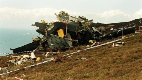 A photograph of the wreck of the Chinook helicopter, which had crashed in Scotland. It is on a large mound of grass, with police tape placed around the crash site.