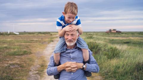 An older man carries a young boy on his shoulders during a walk outside. The relationship is one of grandfather and grandson. 