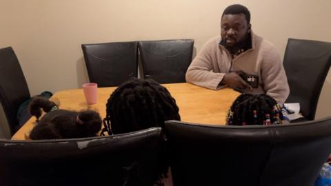 Osei Kwaku sits at the table with his three primary school age daughters. Their backs are to the camera as their parents want to protect them from being identified.