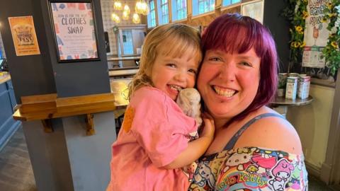 Vikki with purple hair and a Hello Kitty dress smiling while holding a girl in a pink dress in a pub. The girl is holding a soft toy.