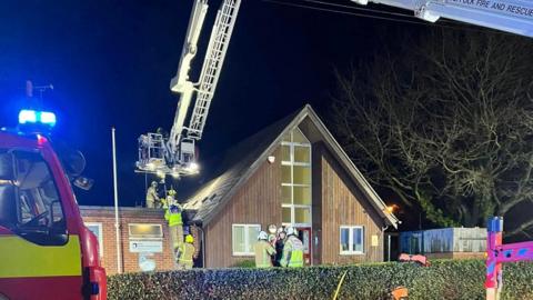 Firefighters can be seen outside a school building working on tackling the fire. Some of the crew stand outside talking while several others can be seen on a ladder leading to the roof.