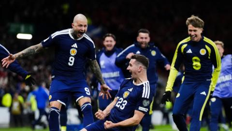 Several Scotland football players celebrate with delight after scoring a goal. One of the team slides on his knees across the turf while others race to hug him.