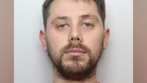 A man with short brown hair and beard in a police mugshot with a grey background