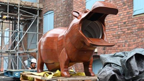 The wooden hippo with its mouth open and perched on a support outside the museum. Behind it is scaffolding and a building.