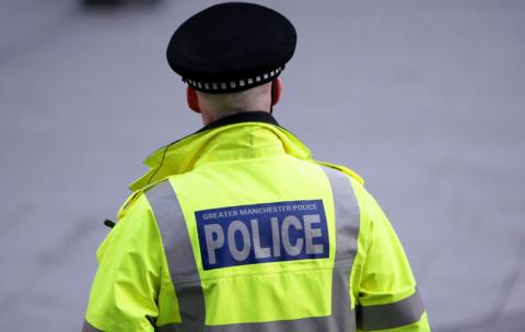 A police officer has his back to the camera. He is wearing a black police hat and is wearing a high-vis jacket with the words 'Greater Manchester Police' on the back.