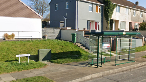 A pavement along a road which has a bus stop on it and a grassy raised verge with a sign that reads UXBRIDGE DR. There is a block of houses behind the bus stop.