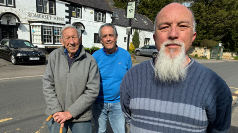 Three middle-aged men stand in front of a white pub. One of them, who has a styled, long white beard, is close to the camera than the other two