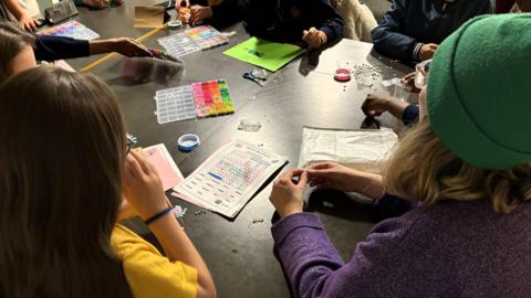 A photo taken above a table. A group of children are sat at the black table. The table is full of paper and crosswords. 