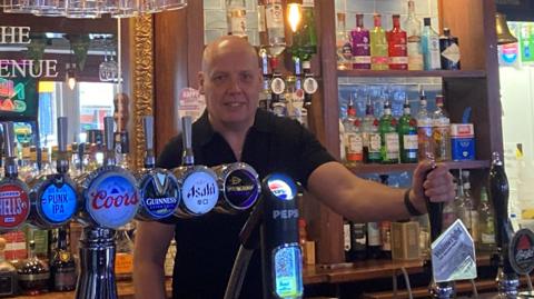 Mr Sharpes stands behind his bar with his hand on the beer pump.