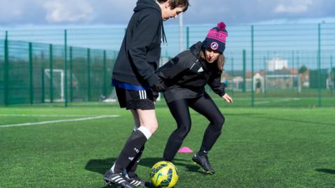 A woman with short dark hair, dark sports top, black shorts, black socks and black football boots running after a yellow and black football with Clare Moss who has long brown hair, a blue and pink hat, green sports bottoms and black football boots. They are on a football pitch surrounded by a green cage.
