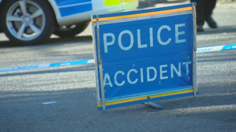 A blue Police Accident sign set up on the road. In the background there is a yellow and blue police car and cordon tape. 