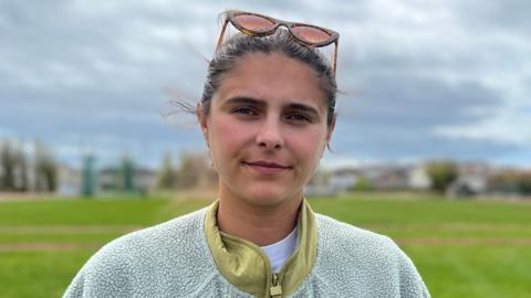 Claudia is standing on a wide grassy field under a cloudy sky. Low buildings and open green space extend into the distance, with sunglasses resting on the her head.