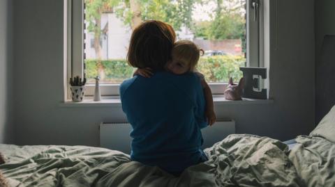 A stock image of a mother and child in a room, with a bed, a large window, with an E to the right. The mother is holding her child on one shoulder and she is wearing a blue top.