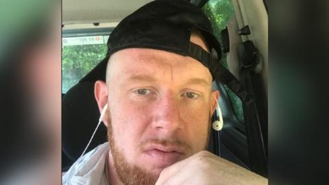 A man with a beard wearing a black baseball hat and earphones. 