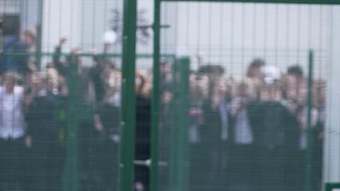 View of the protest children wave pieces of paper as they stand behind a green wire fence. The picture has been blurred to protect their identities