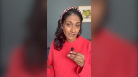 A woman wearing a red jumper holds a small microphone and looks directly at the camera