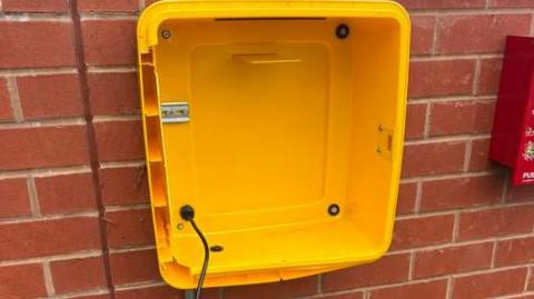 A yellow defibrillator housing on a red-bricked wall. The equipment has been removed and there is a black cable hanging down.