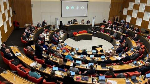 The council chamber. Seating is arranged in curved tiers. The council members sit with computers in front of them.