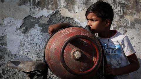 A boy stands next to an empty LPG cylinder tied to a bicycle as he waits outside a gas agency in India