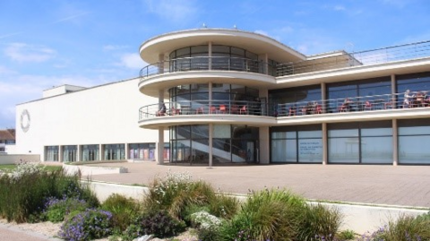 A building with a large round section and glass panels on the front of the building. The building is set against a blue sky and there are plants in the foreground.