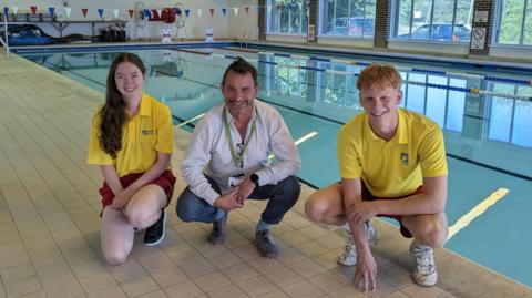 Three people squatting by a swimming pool. 