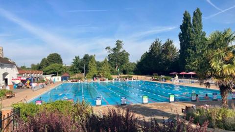 Guildford Lido, an outdoor pool, is seen under a blue sky and it is surrounded by trees. The water is blue and there are lane dividers out and some swimmers swimming lengths.