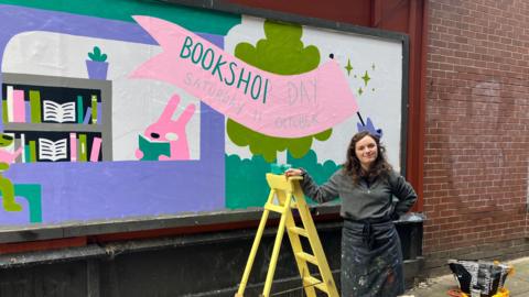 A person stands beside a yellow ladder in front of a vibrant mural promoting 'Bookshop Day' on Saturday 11 October. The mural features colourful illustrations of bookshelves, books, and cartoon animals, with a prominent pink banner displaying the event details. Artist Bethan Woollvin is dressed in a dark apron and holding paint supplies. Painting tools and materials are scattered on the ground nearby.
