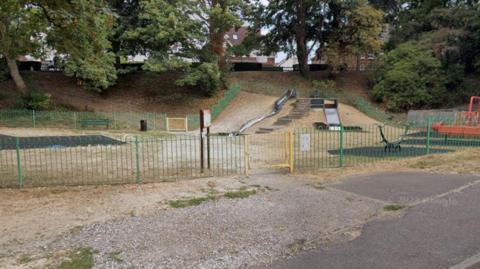 The image shows a playground that contains a slide, a seesaw and some swings. Behind the park are trees and some houses that can be seen behind them.