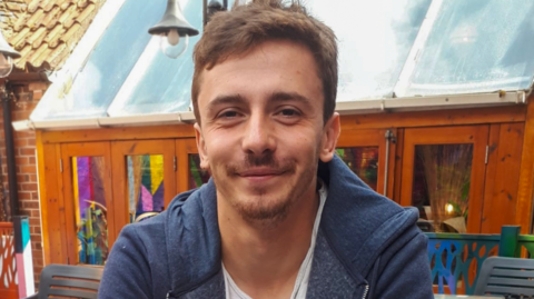A smiling man sat down in an outdoor location. He has brown hair and a short beard and is wearing a blue hooded top.