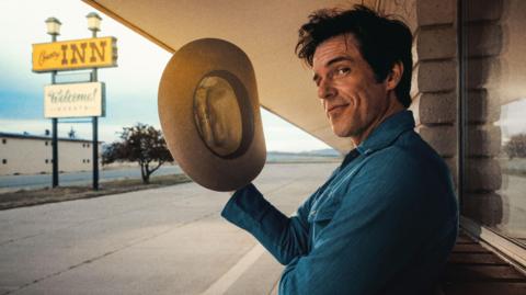 Brandon Flowers holds a stetson in his right hand and gives a wry look to the camera as he poses outside a stereotypical American motel