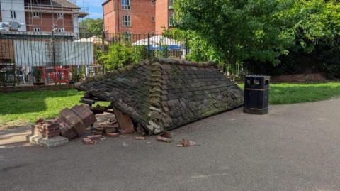 Bricks and a large tile roof of a historic outdoor shelter. The structure has collapsed next to a black public waste win on a concrete surface. In the background are trees and grass, with residential properties beyond them.
