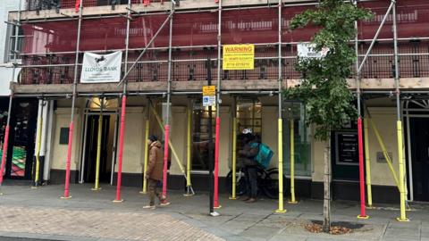 A bank surrounded by scaffolding and safety netting. The "Lloyds Bank" branding is partially visible behind the scaffolding.