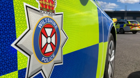 Stock image of a Wiltshire Police car. It features a grey, yellow and blue logo which reads 'Wiltshire Police' on the side of a yellow and blue vehicle.