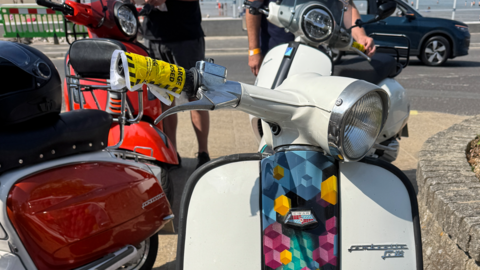 Close-up of a white scooter with a multicoloured decorative strip down the centre of the front faring and a yellow parking fine taped around one of the handlebars. Three other scooters are parked in the background.