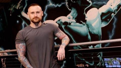 A man with short brown hair, a beard, tattoos on his arms and neck, and wearing a brown shirt, leans on a barrier. Behind him is an artwork depicting Arnold Schwarzenegger. 