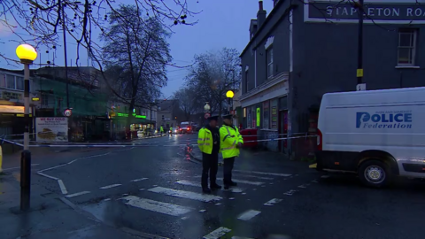Two police officers stand on a zebra crossing with their backs facing a cordon at a murder investigation scene on a road lined with shops in Bristol. It is a grey and damp morning, which is still quite dark. A police federation van is parked beside them.
