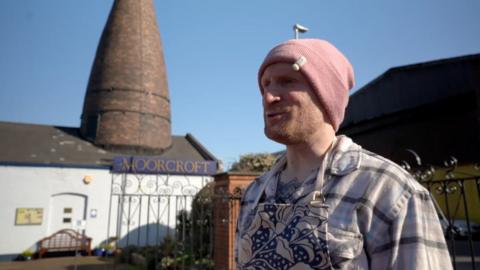 A man in a pink hoodie stood in front of a brown bottle kiln. He's wearing a short and blue and white apron